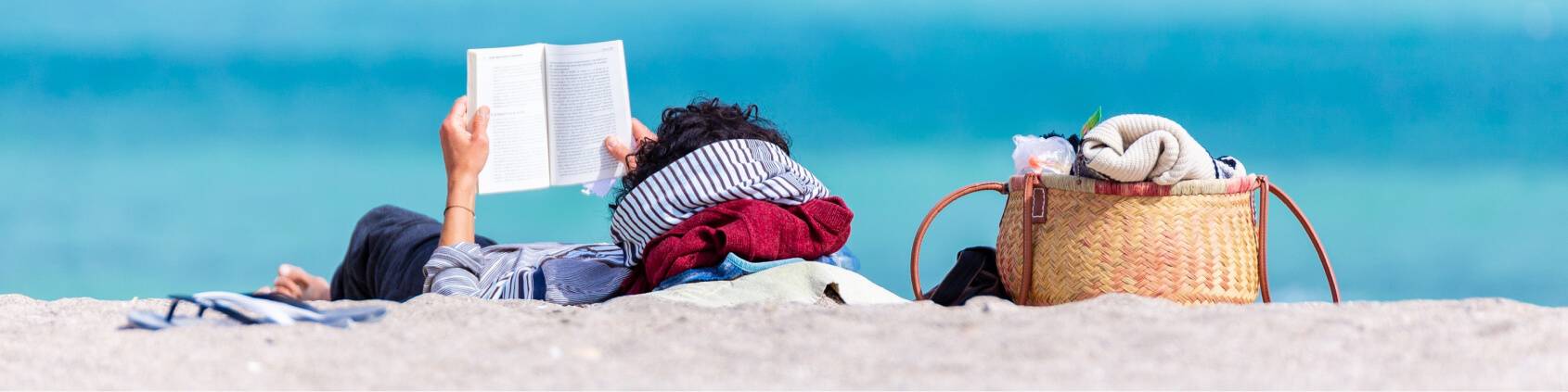 woman lays on the sand at the beach reading a book. She looks relaxed.